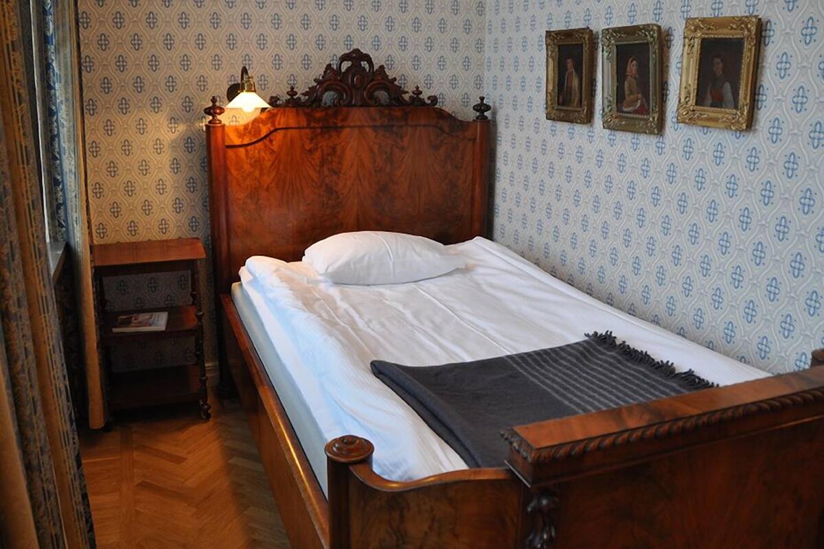 A wooden bed with white linens and a dark throw blanket, a nightstand with a lamp, and patterned wallpaper with three framed pictures on the wall.