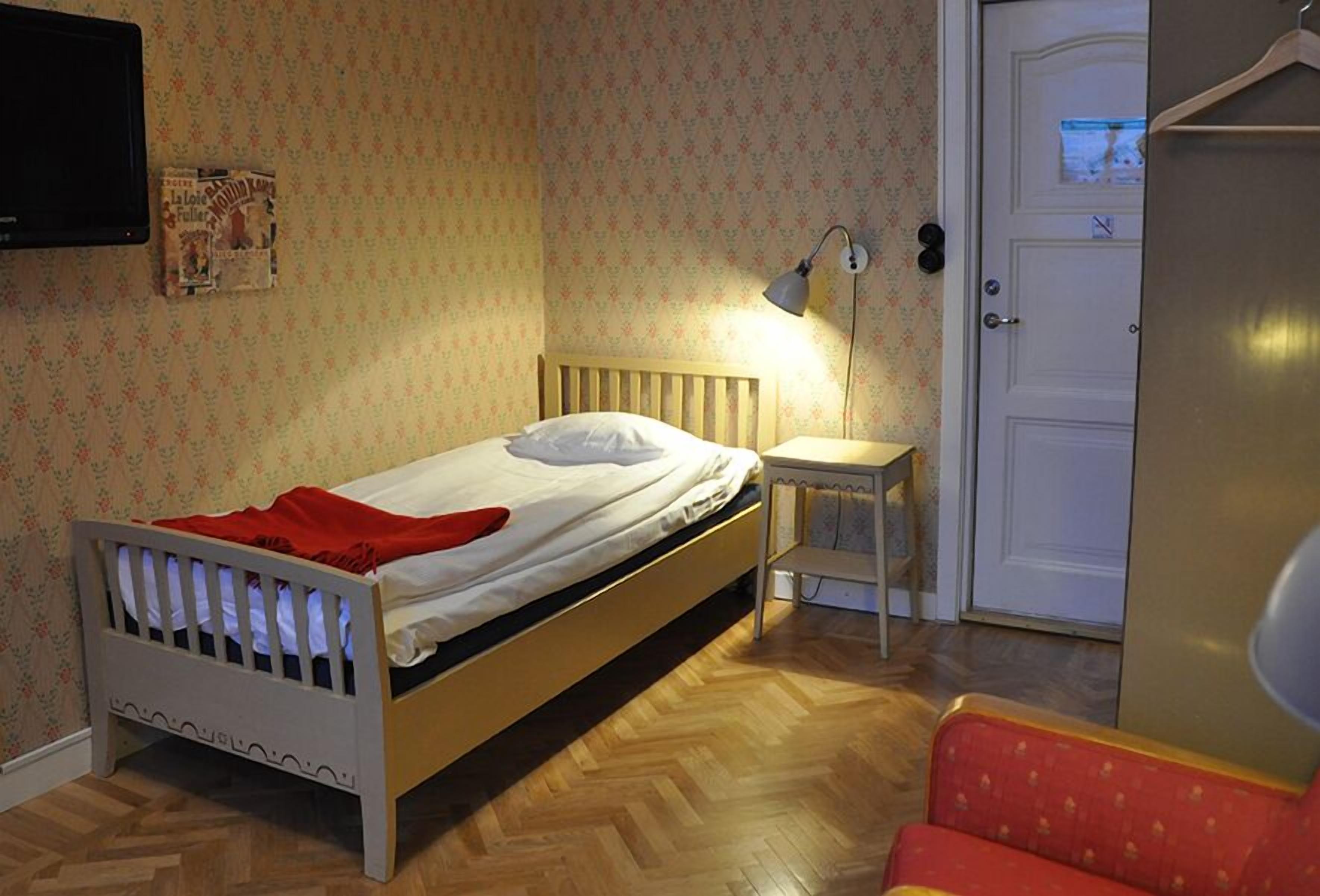 Un dormitorio con cama de madera, una mesita de noche pequeña, un televisor montado en la pared y una puerta con manija.