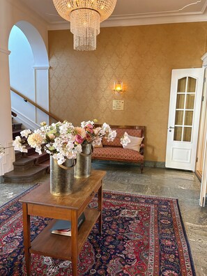 A lobby with a chandelier, a reception desk with vases of flowers, a patterned rug, a sofa with cushions, and a door with a sign. - Laholms Stadshotell (Laholm)