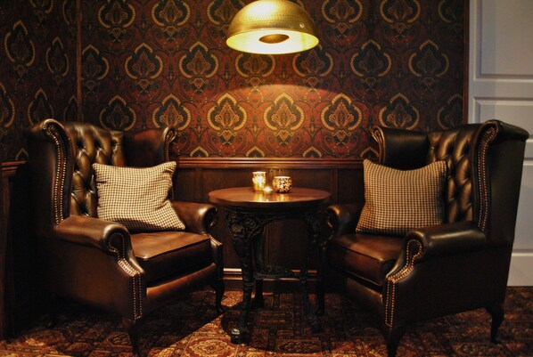 Een kamer met twee leren fauteuils, een klein rond tafeltje met een kaars, en behangpapier met dessin.