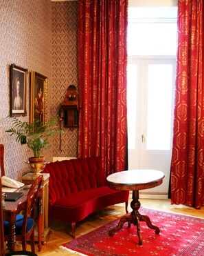 Een kamer met een rode bank, een ronde tafel, een houten stoel, een potplant en een lijst met een afbeelding aan de muur.