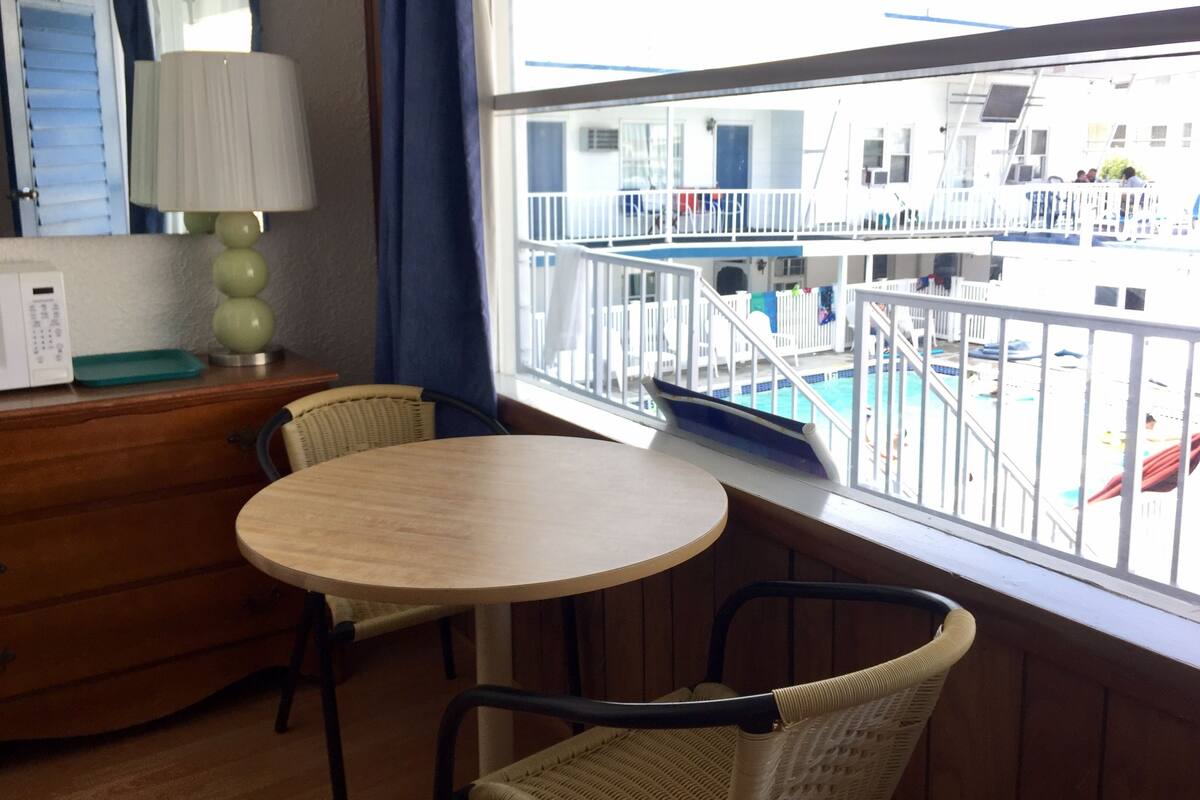 Una habitación con una mesa redonda de madera y dos sillas de mimbre, un aparador de madera, una lámpara y una ventana con vistas a una piscina y un edificio.