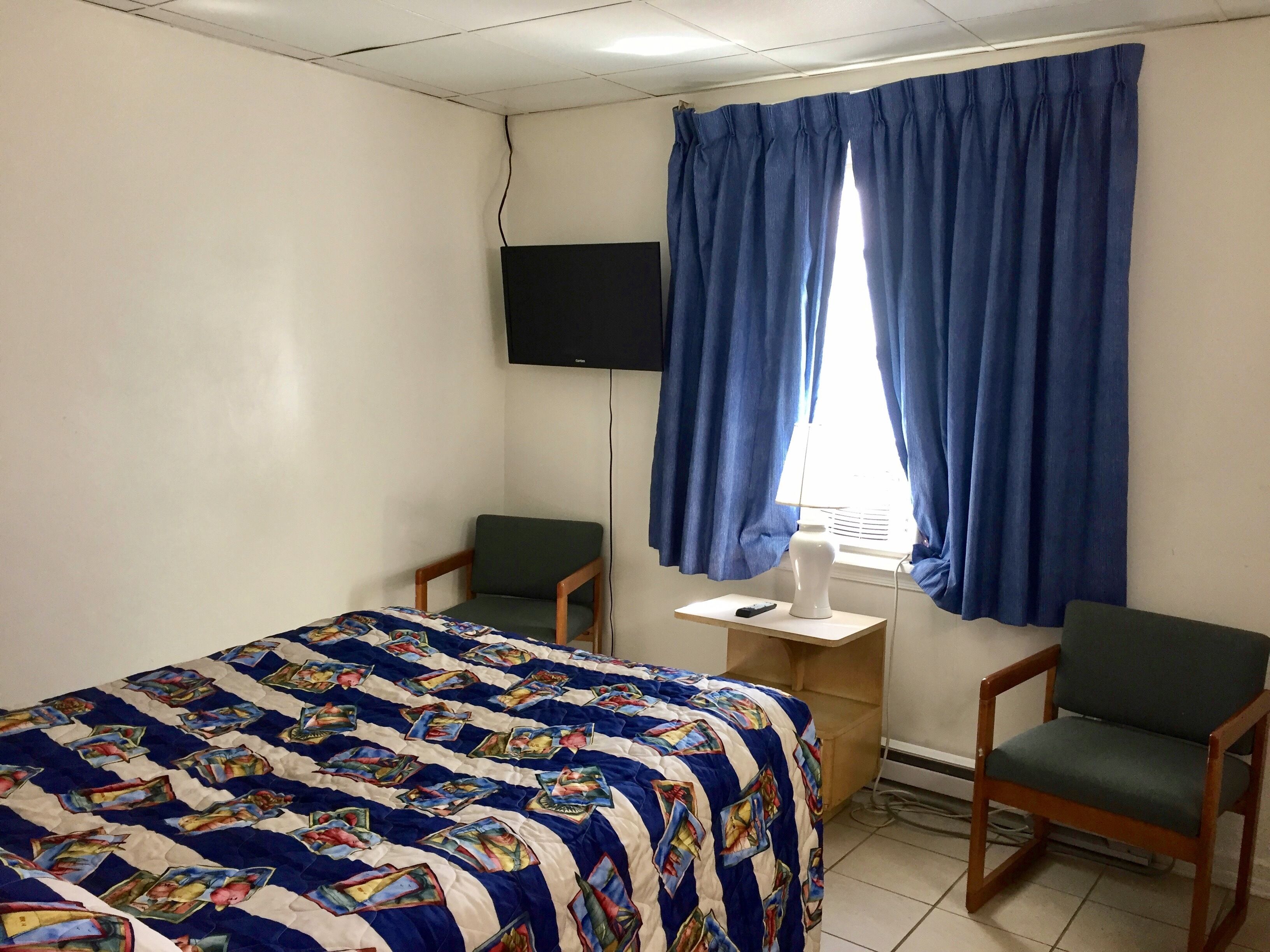 Una habitación de hotel con cama, dos sillas, televisor y ventana con cortinas.