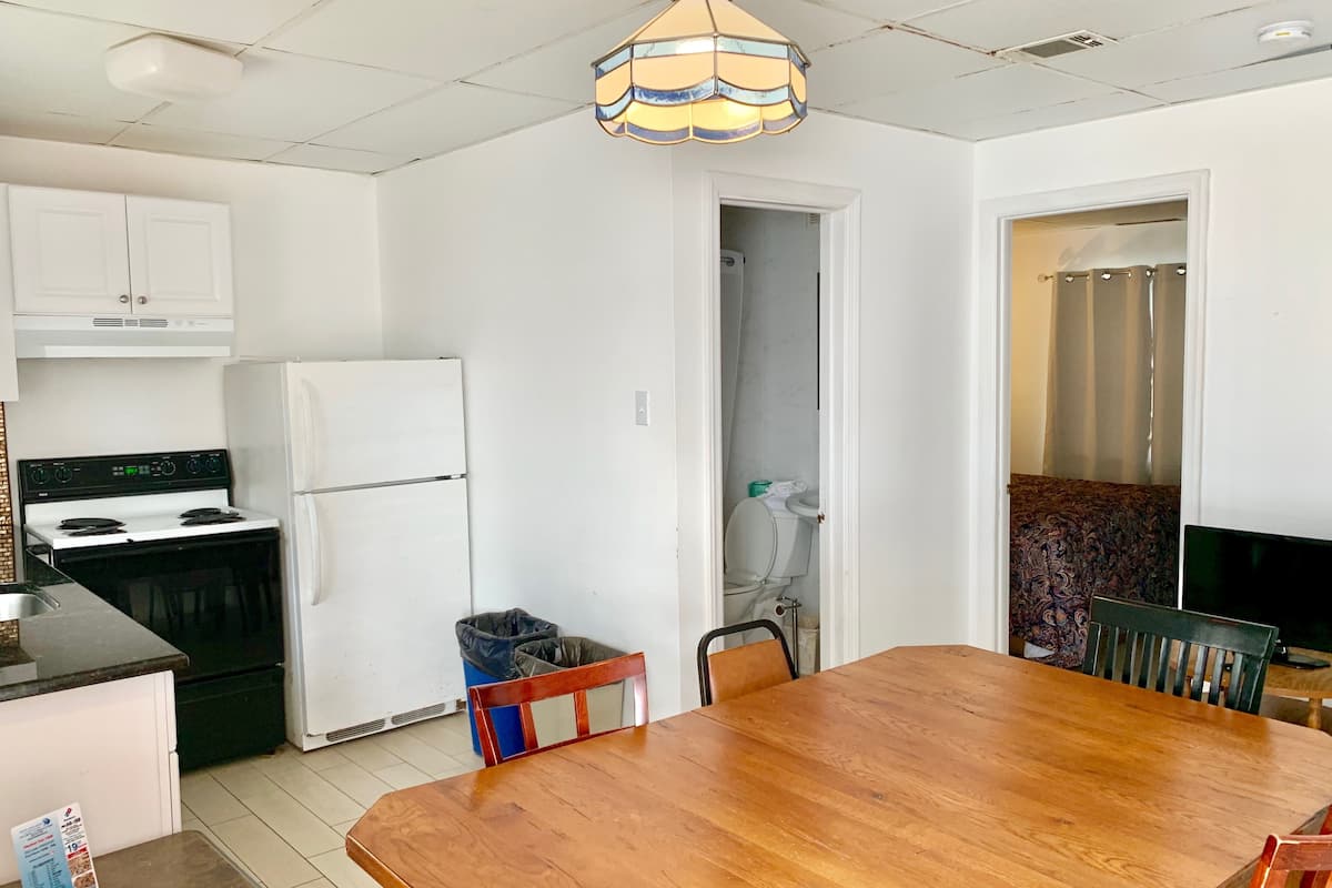 Una cocina con armarios blancos, una estufa y un refrigerador. Hay una mesa de comedor con sillas y un bote de basura.