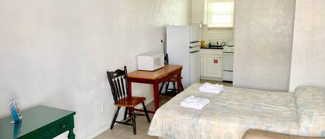 A small, clean room with a bed, a wooden chair, a small table, and a kitchenette in the background.