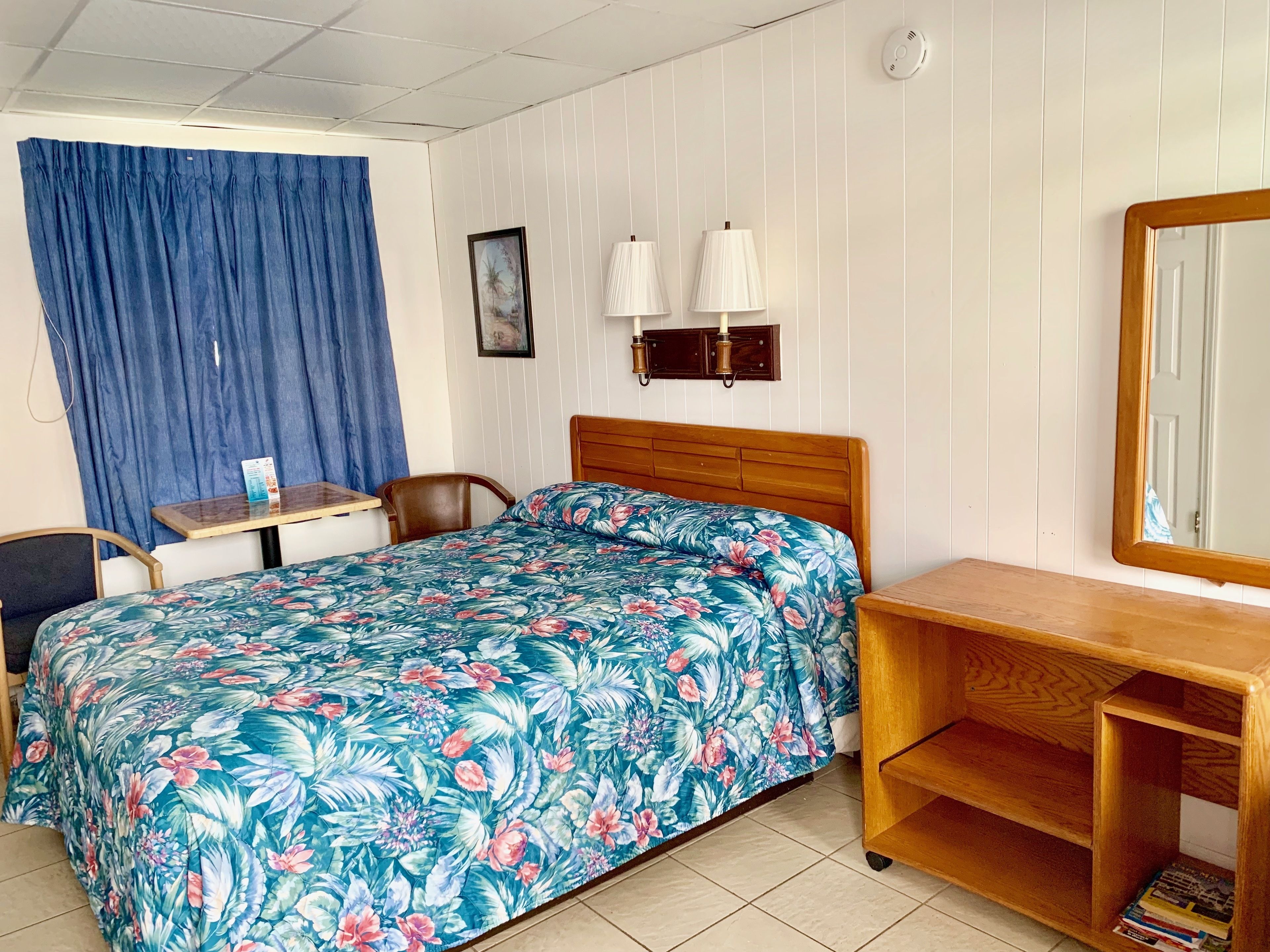 A hotel room with a bed, a desk, a chair, a wooden nightstand, a mirror, and a blue curtain.