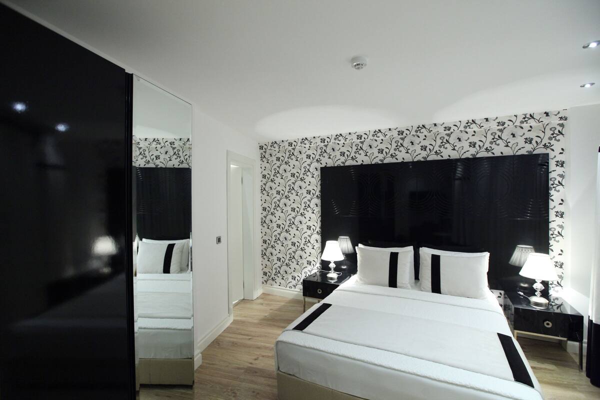 Et moderne hotelværelse med en stor seng, to sengeborde med lamper, et stort spejl og et blomstret tapetdesign.