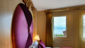 A hotel room with a large bed, a view of the sea through a window, and a lamp on the bedside table.