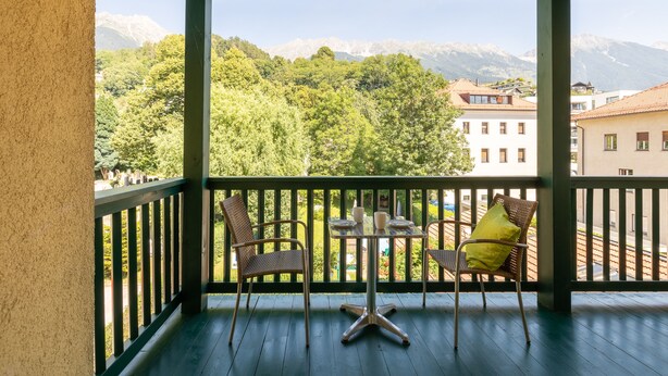 A balcony with wicker chairs and a table, overlooking a mountainous landscape and buildings.