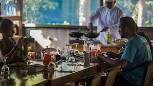 A restaurant setting with a chef preparing food at the counter, a table with various dishes and drinks, and a view of greenery outside. - Muyuna Lodge (Fernando Lores)