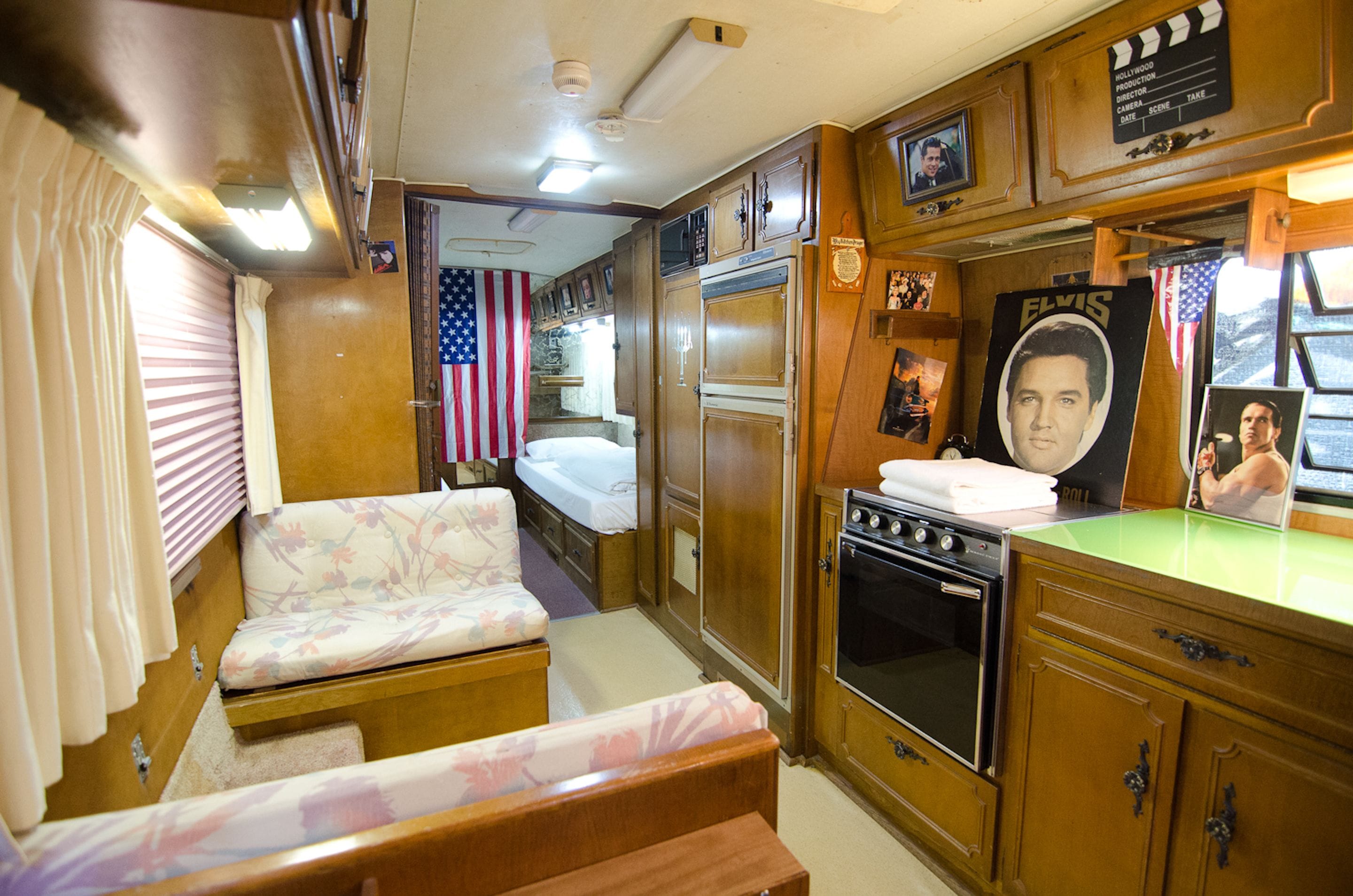 A compact RV interior with a bed, kitchenette, and a poster of Elvis Presley.