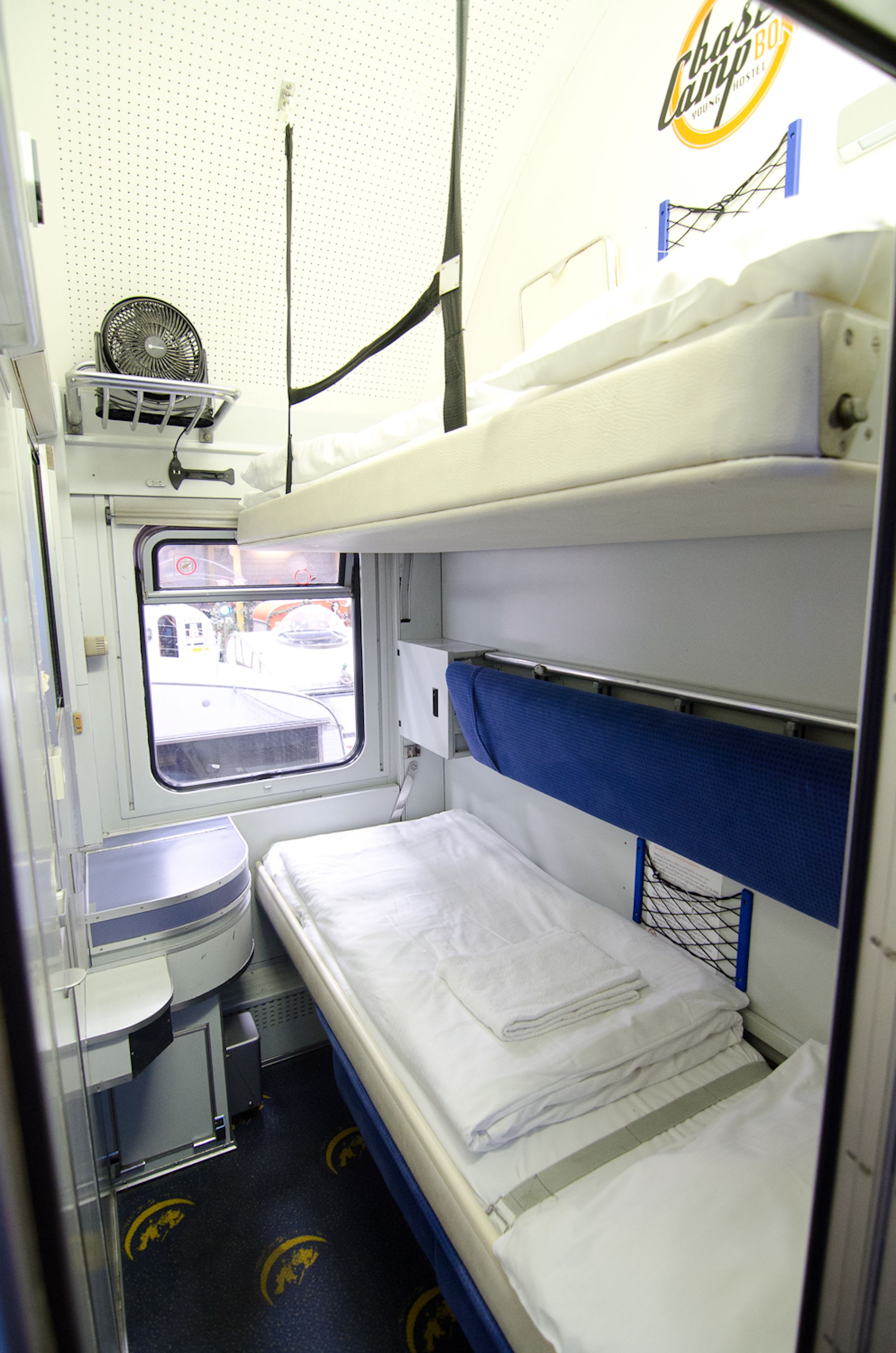 A train compartment with a bunk bed, a small table, and a window providing a view of the outside.