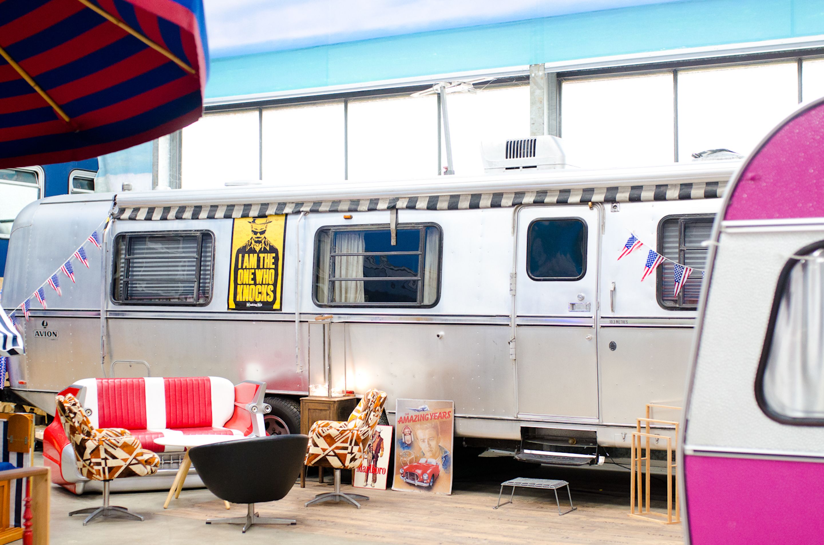Una caravana vintage con un toldo a rayas, un sofá rojo y blanco, y una silla negra.