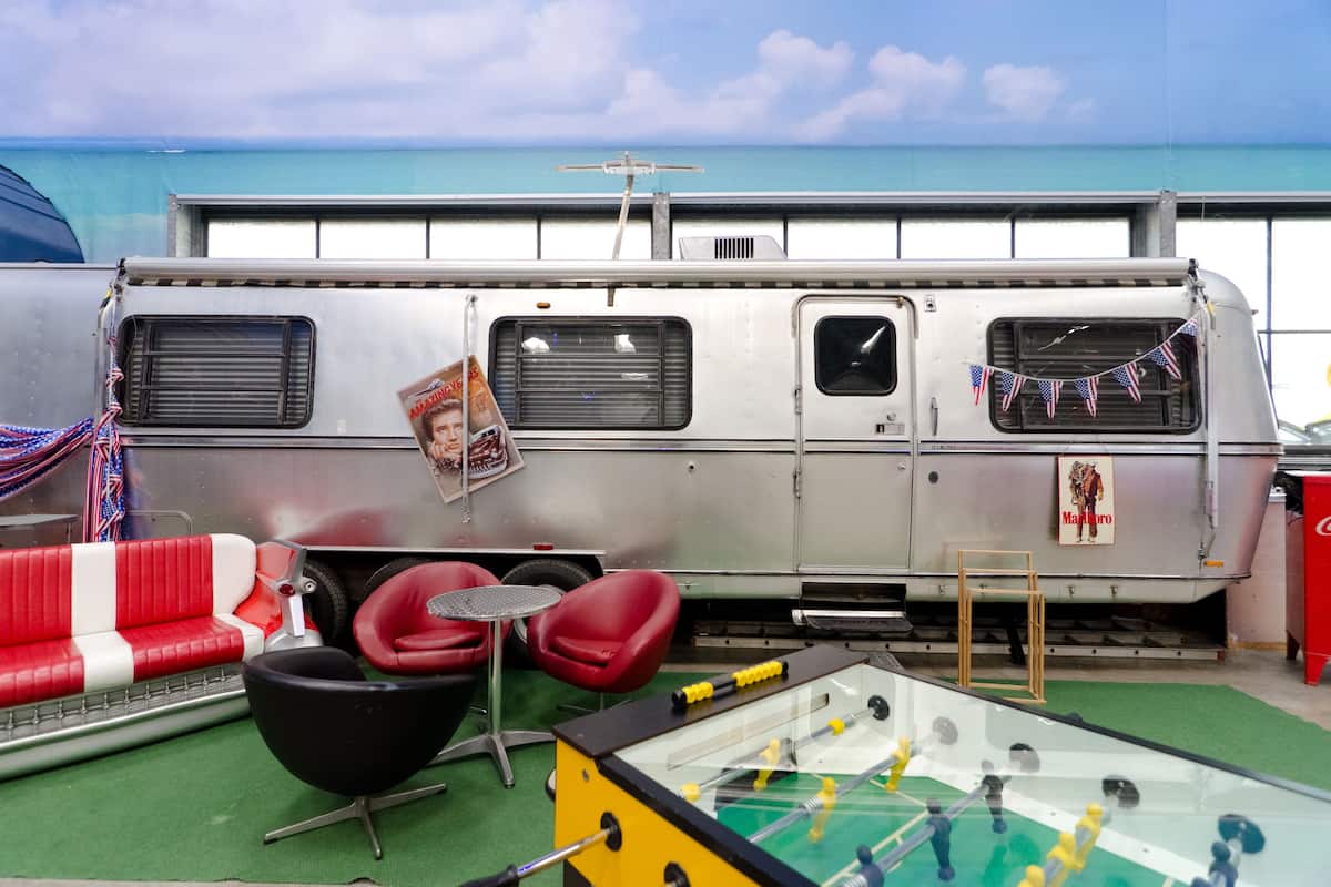 A retro-themed room with a vintage camper van, red and white striped sofa, red chairs, and a foosball table.