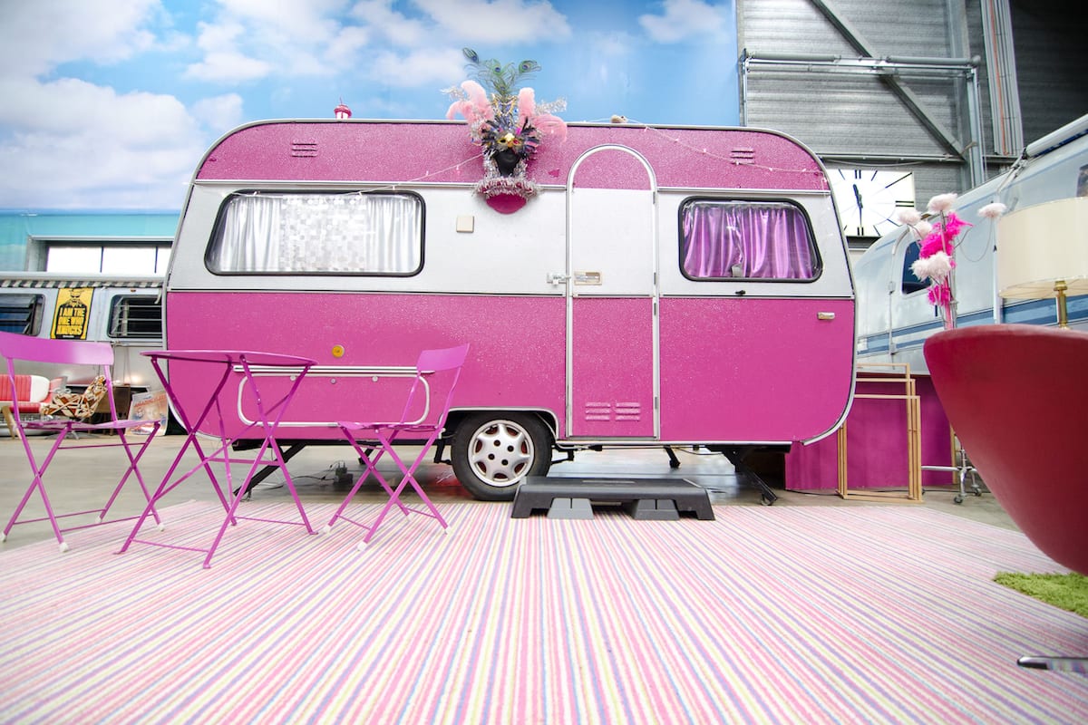 A pink RV with a small table and chairs in front, a step leading up to the entrance, and a decorative flower arrangement on the side.