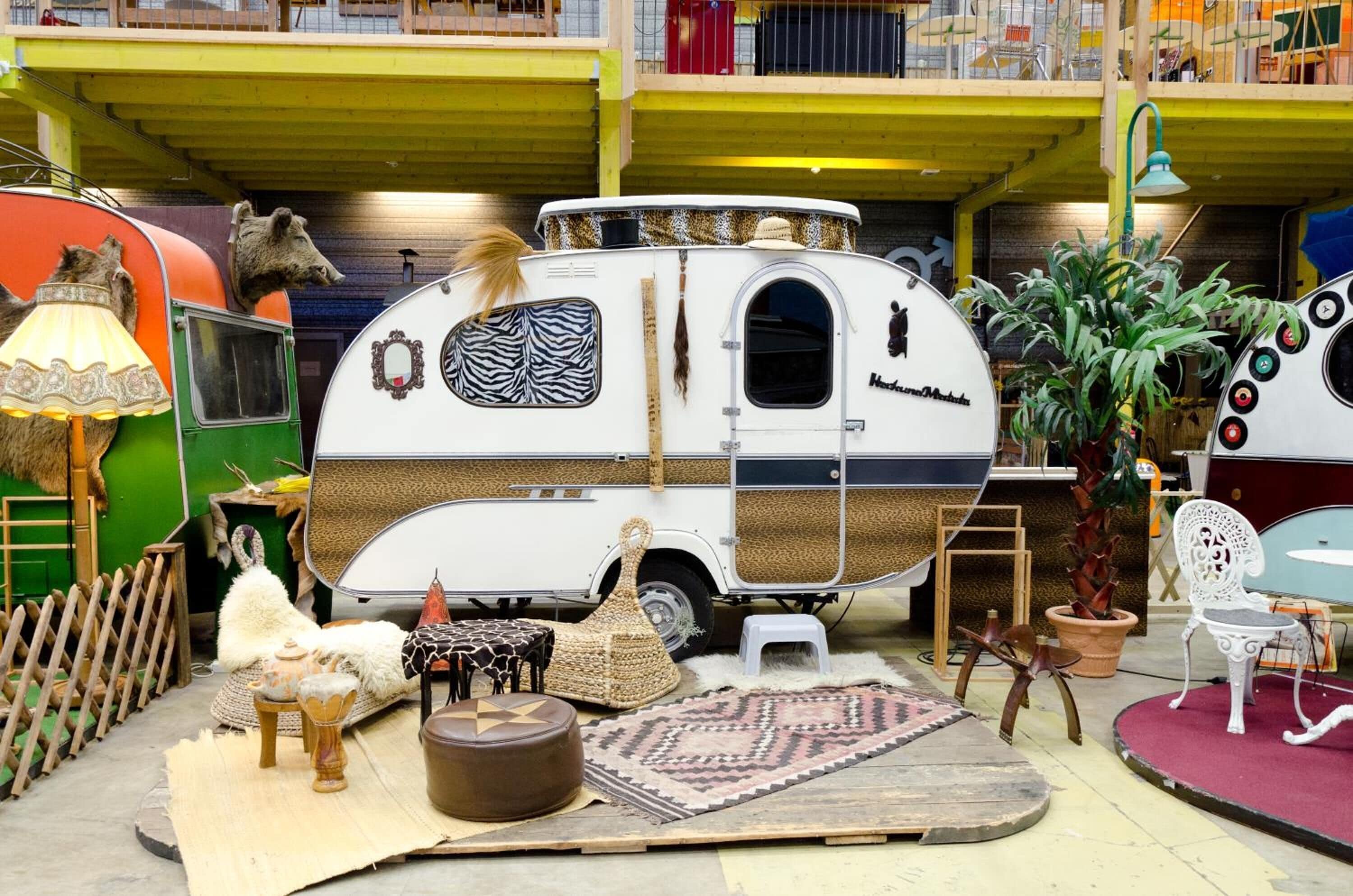 A vintage camper with a zebra-patterned side panel, surrounded by rustic decor including a woven basket, a small stool, and a patterned rug.