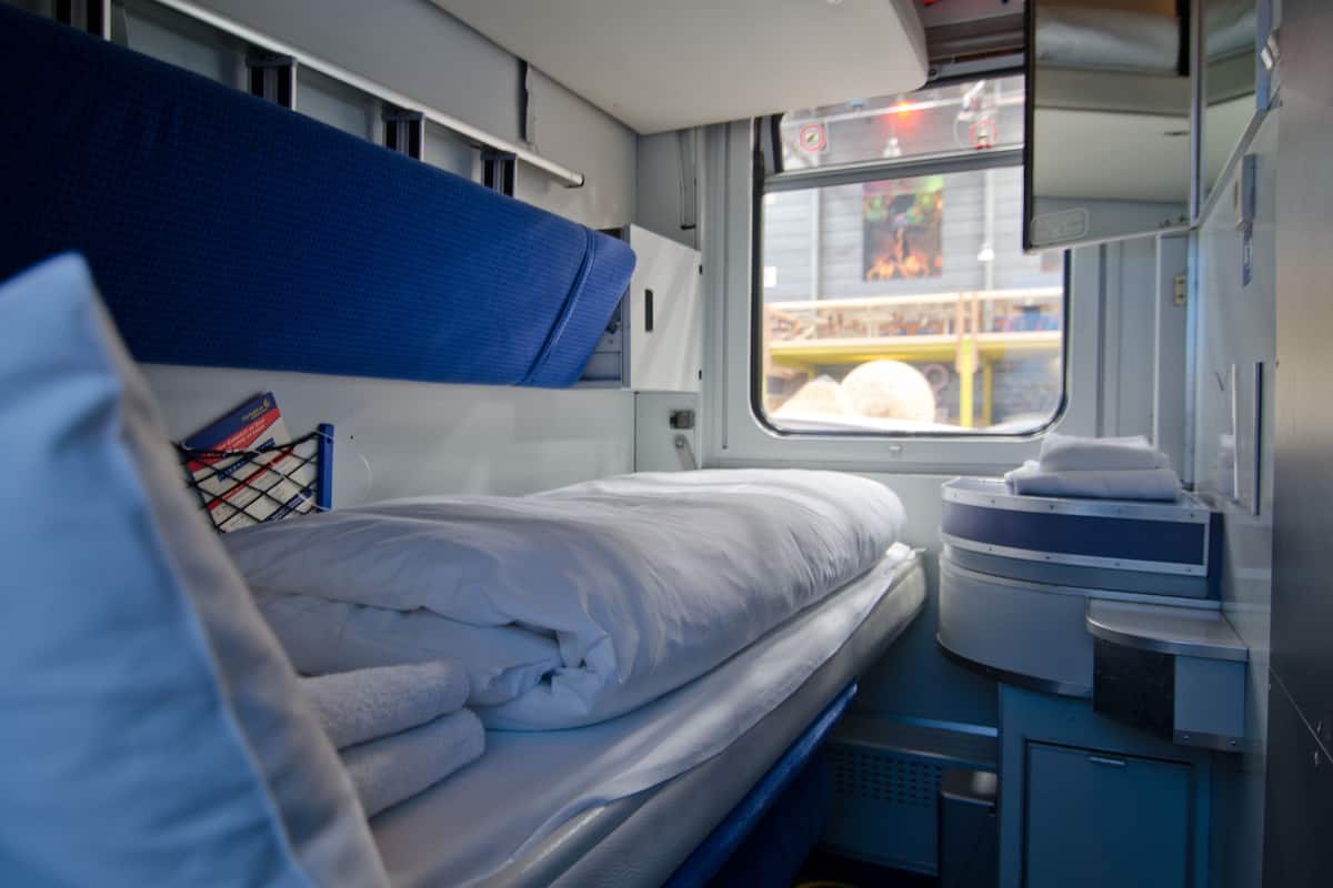 A train compartment with a bunk bed, a small table with a book, and a window with a view of the outside.