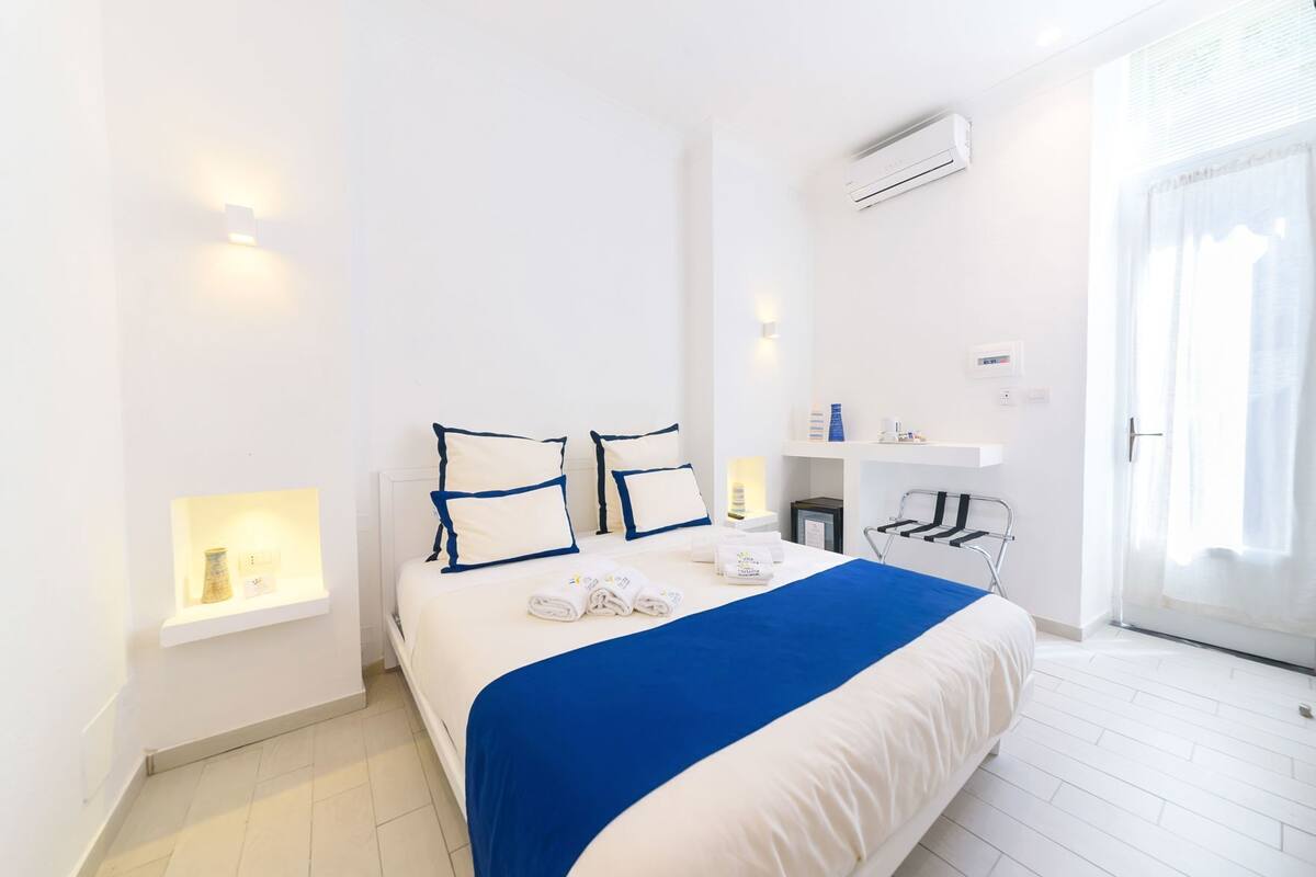 A hotel room with a bed, bedside tables, an air conditioning unit, a bedside lamp, and a window with curtains.