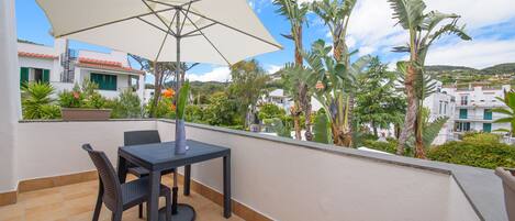 A balcony with a table and chairs, a large umbrella, and a view of buildings and palm trees.