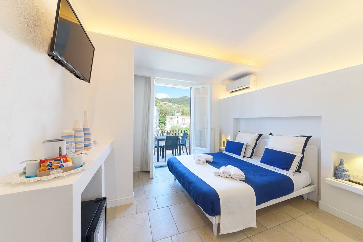 A hotel room with a bed, a TV mounted on the wall, a small table with toiletries, and a view of the outside.