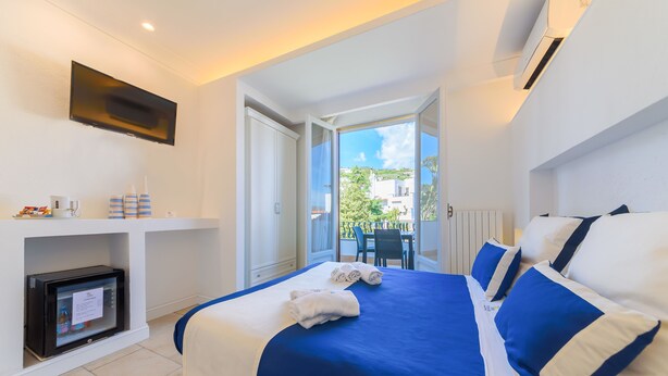 A hotel room with a bed, a TV, a mini-fridge, and a balcony with a table and chairs. - Villa Fortuna Holiday Resort (Ischia)