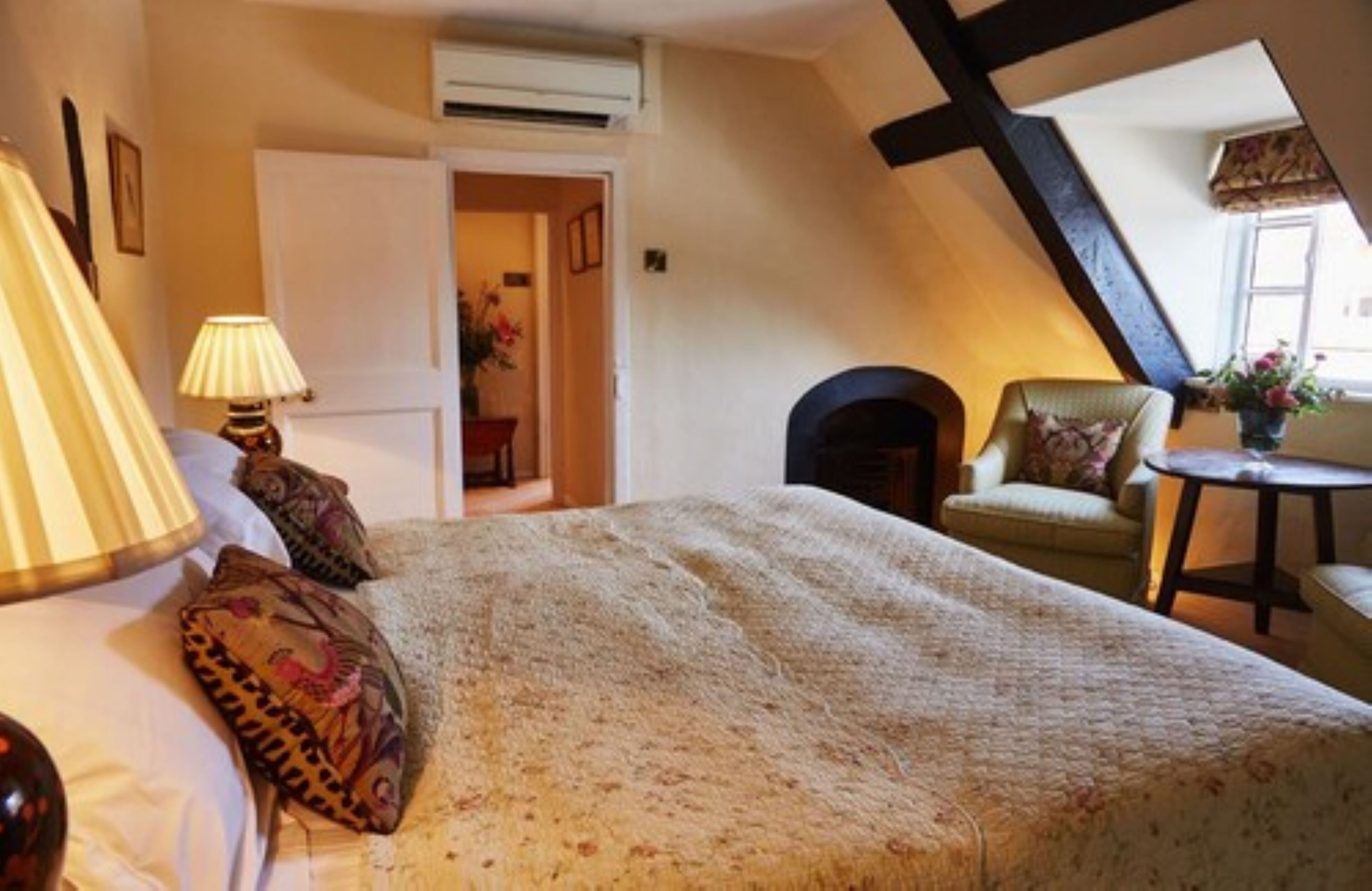 A bedroom with a large bed, a chair, a small table, and a lamp. There is a door leading to another room.