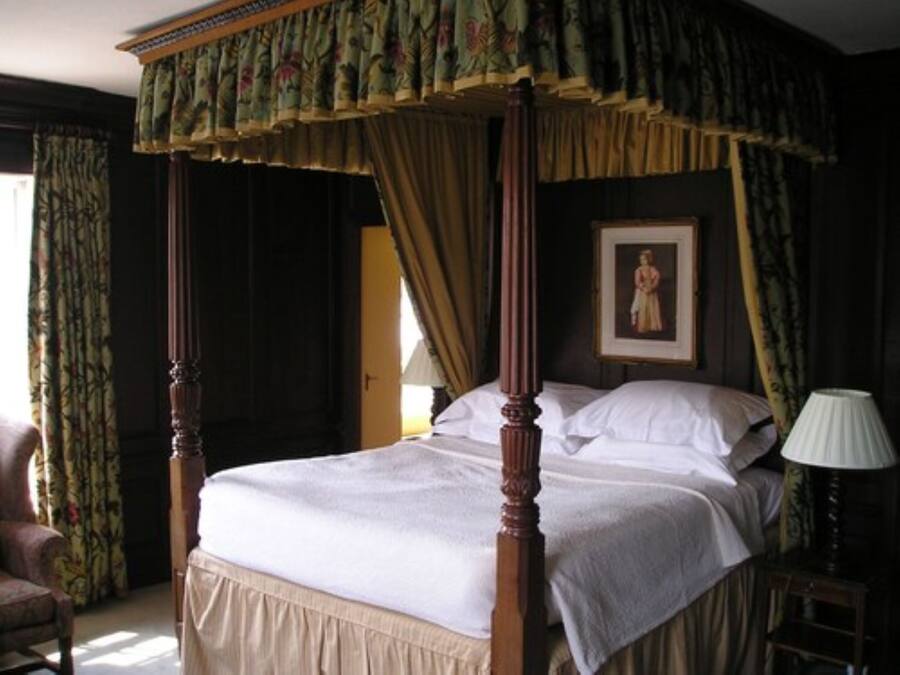 Una cama con dosel y cuatro columnas, con una colcha blanca, un cuadro en la pared y una lámpara en una mesita de noche.