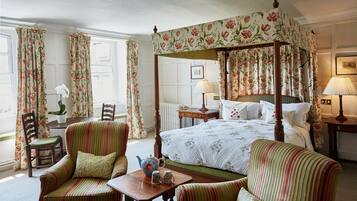 A four-poster canopy bed with floral curtains, a sitting area with striped armchairs, a small table with a teapot, and a view of the room's white walls and window.