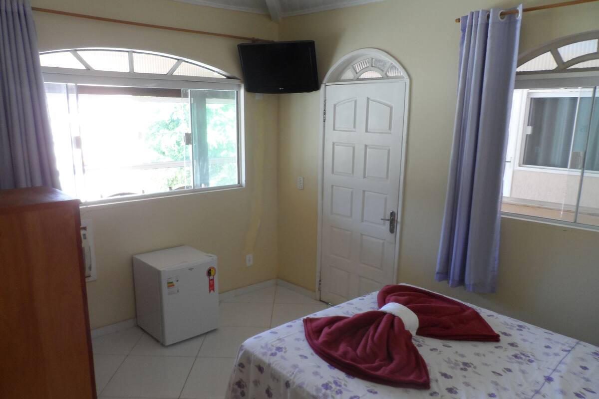Una habitación con cama, televisor montado en la pared, refrigerador pequeño y ventana con cortinas.