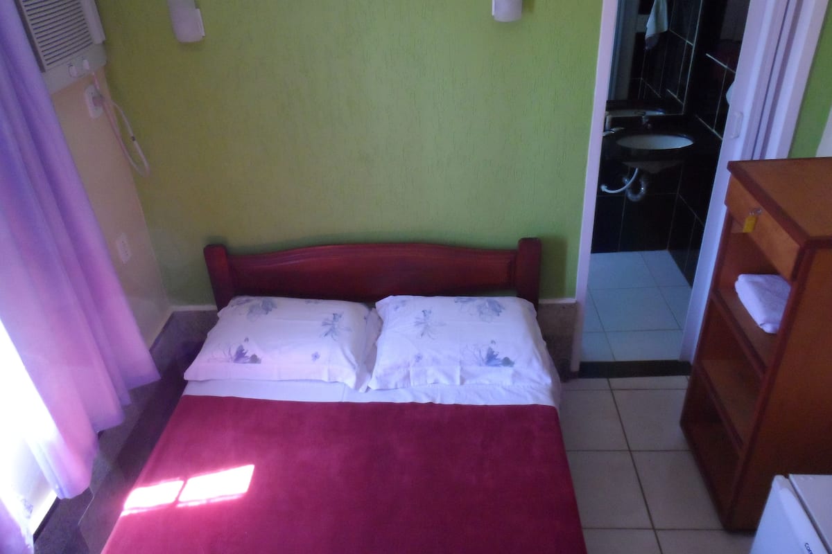 Una habitación con una cama, un aparador de madera, un baño visible a través de una puerta abierta y un equipo de aire acondicionado instalado en la pared.