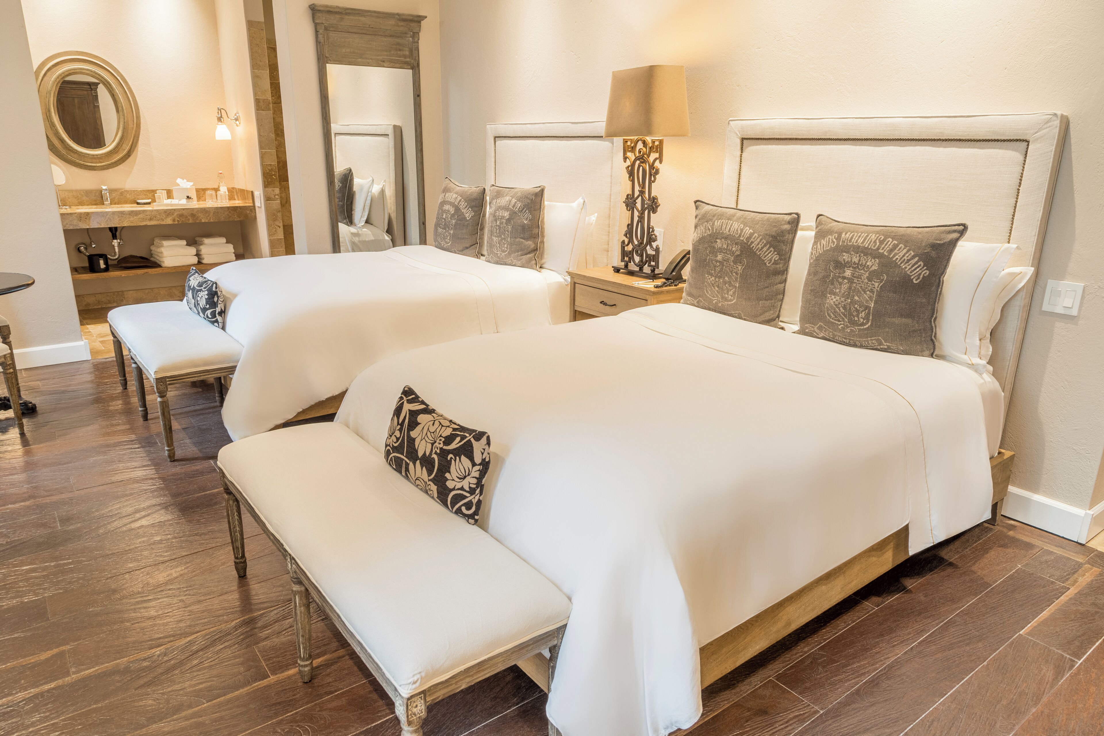 Una camera d'albergo con due letti, una panca, un comò con uno specchio e un comodino con una lampada.