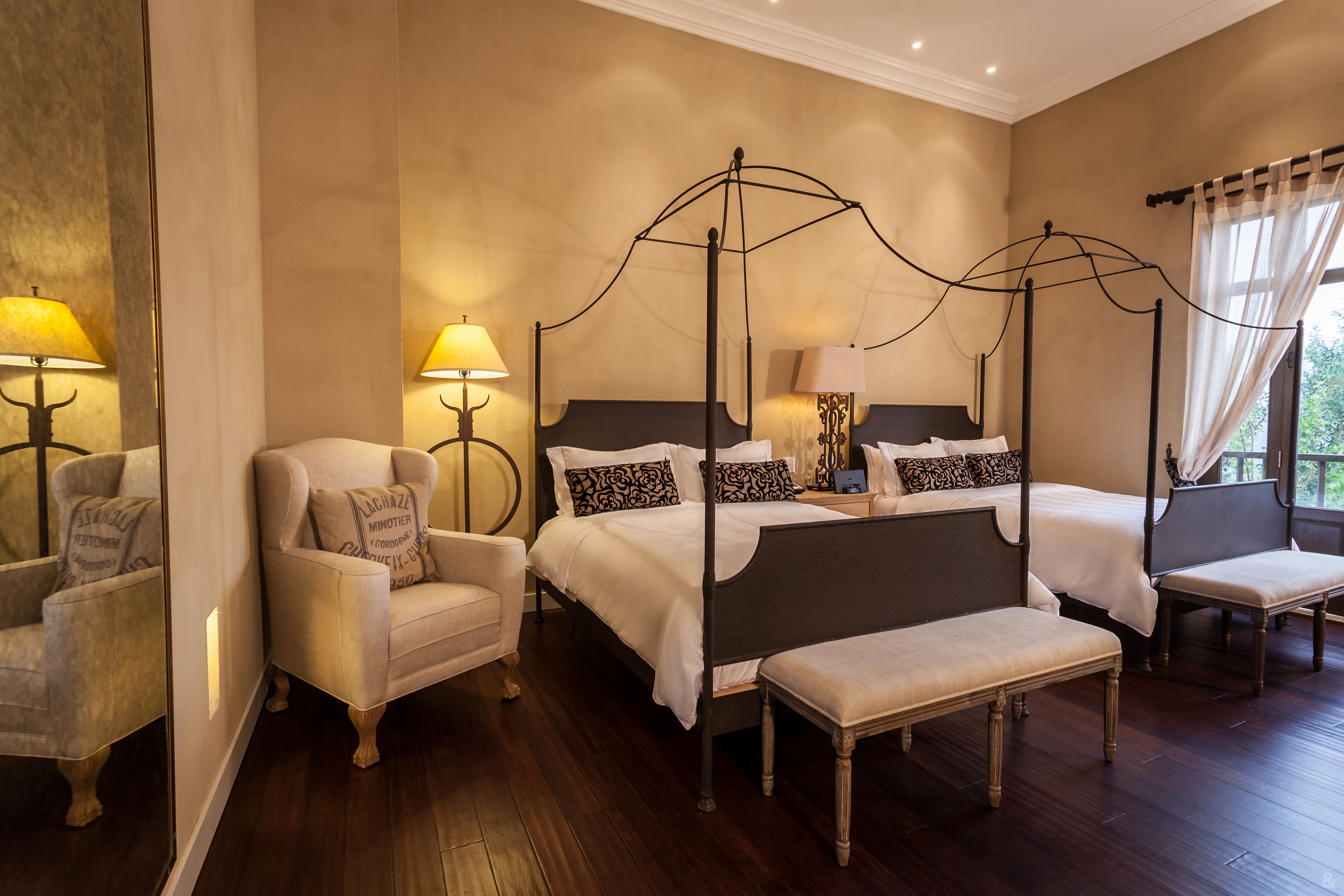 A four-poster bed with a dark headboard, two bedside tables with lamps, a chair, and a bench.