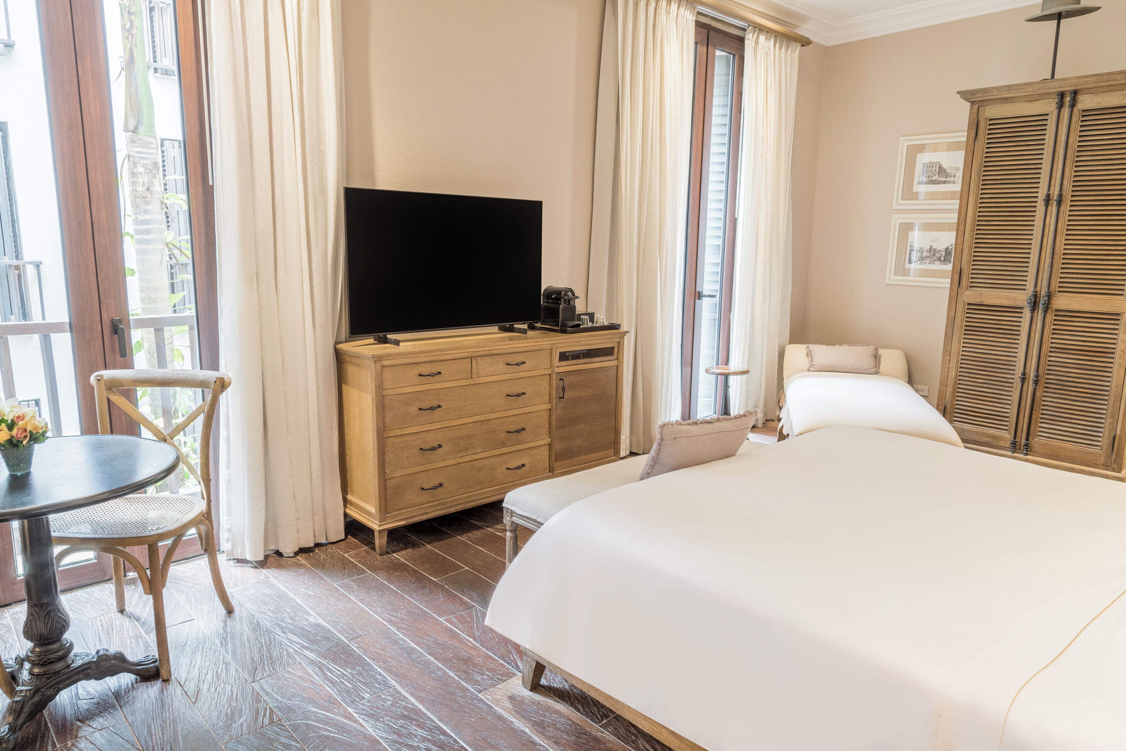 A hotel room with a large bed, a wooden dresser with a flat-screen TV, a small round table with a chair, and a window with curtains.