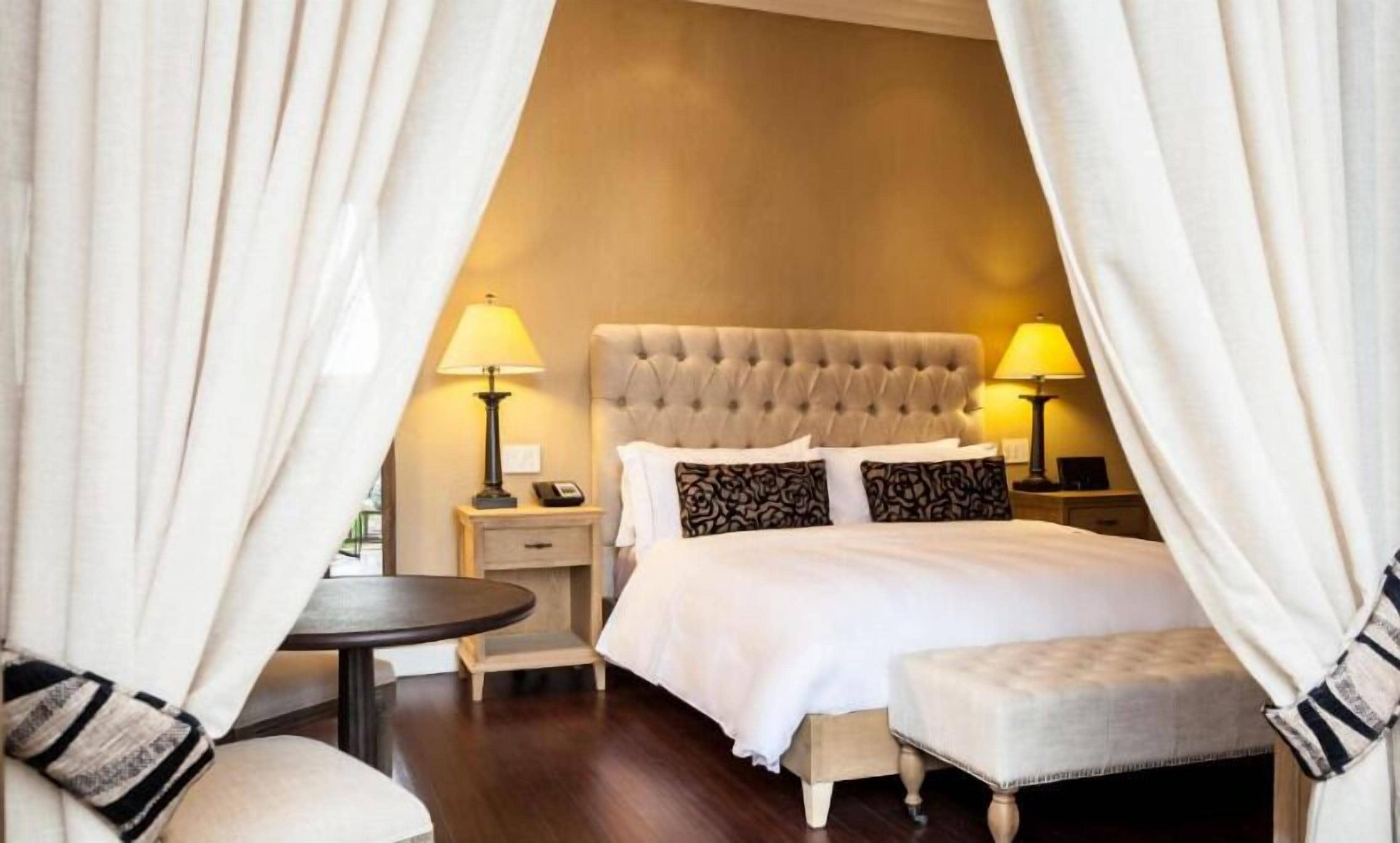 Una habitación de hotel con un cabecero capitonado, una cama con ropa de cama blanca, dos lámparas de noche, una mesita de noche, una pequeña mesa redonda y un reposapiés.