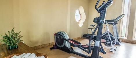 Un gimnasio en casa con dos bicicletas estáticas, una máquina de remo y una mesa de madera con toallas enrolladas.