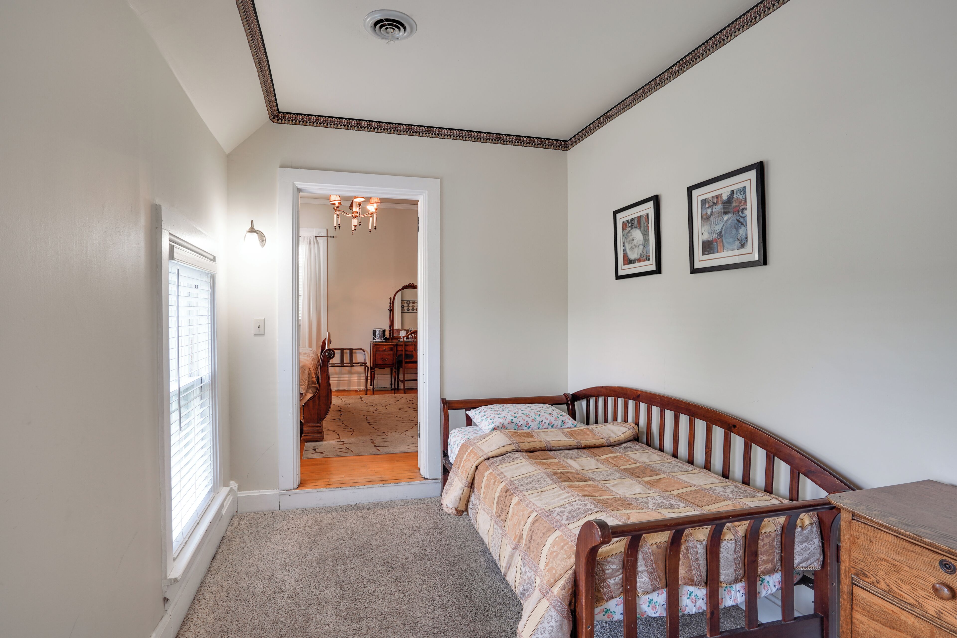 A bedroom with a wooden bed, a nightstand, a door leading to a hallway, and a ceiling with a decorative border.