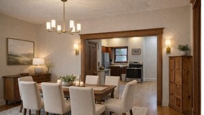 A dining area with a wooden table and chairs, a chandelier, and a view into the kitchen. - White Rose Inns (Wisconsin Dells)