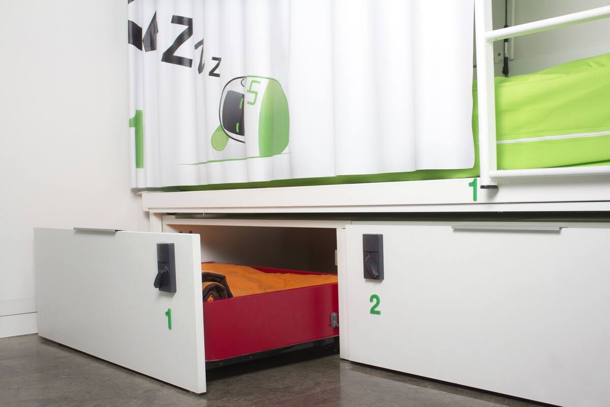 Uma cama beliche com colchão verde, parede branca com logotipo verde e branco, e um compartimento de armazenamento vermelho.