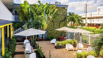 Un espace extérieur aménagé avec des chaises blanches et des parasols, entouré de plantes et agrémenté d’un mur décoratif portant une inscription.
