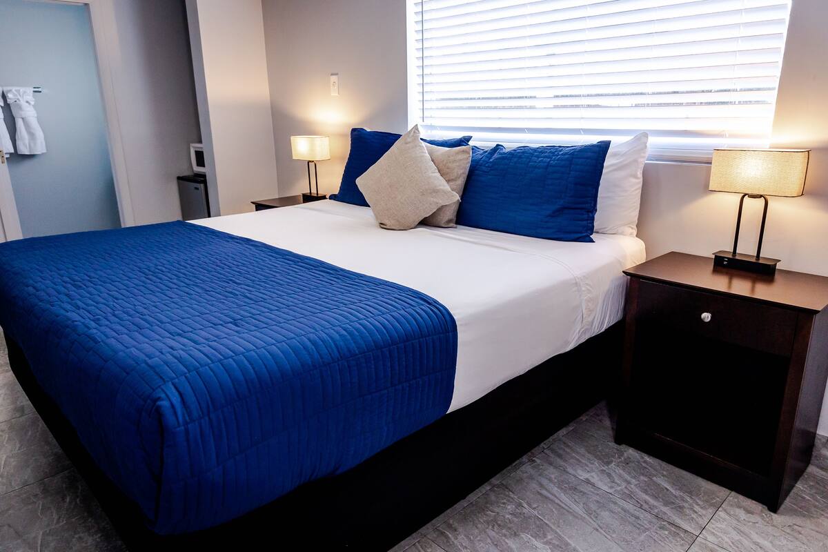 Un lit bien fait, avec une couette bleue, du linge de lit blanc et plusieurs oreillers, dans une chambre d’hôtel offrant une vue sur la ville par la fenêtre.