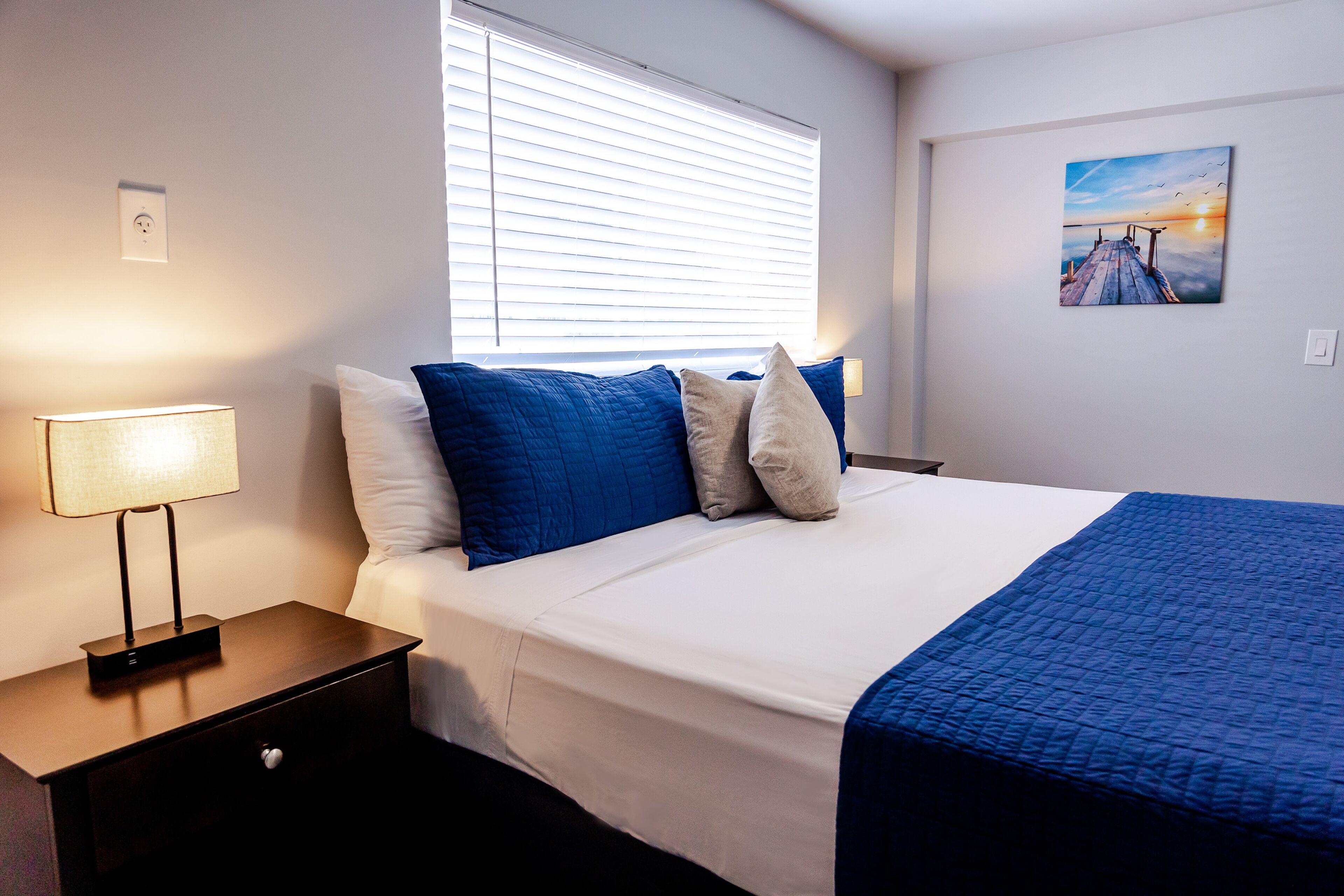 A neatly made bed with blue and white bedding, a bedside lamp, and a painting of a sunset on the wall.
