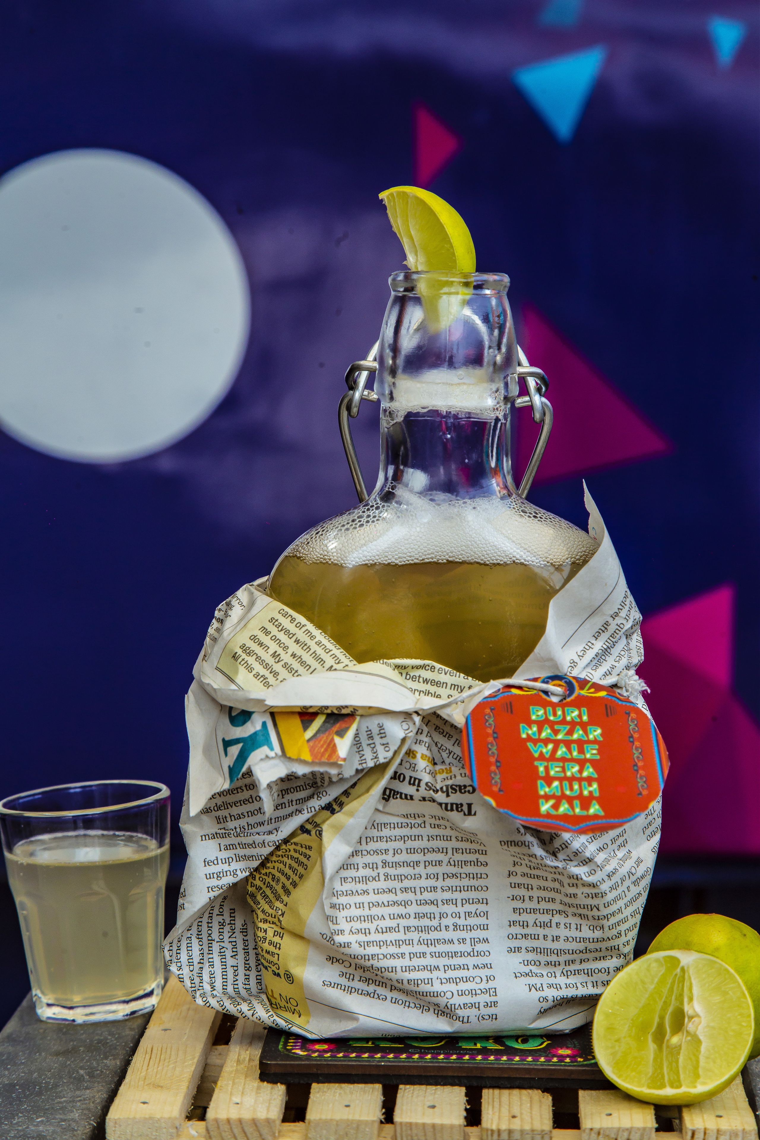 A glass bottle with a lemon slice on top, a glass of liquid beside it, and a lime half on a wooden surface.