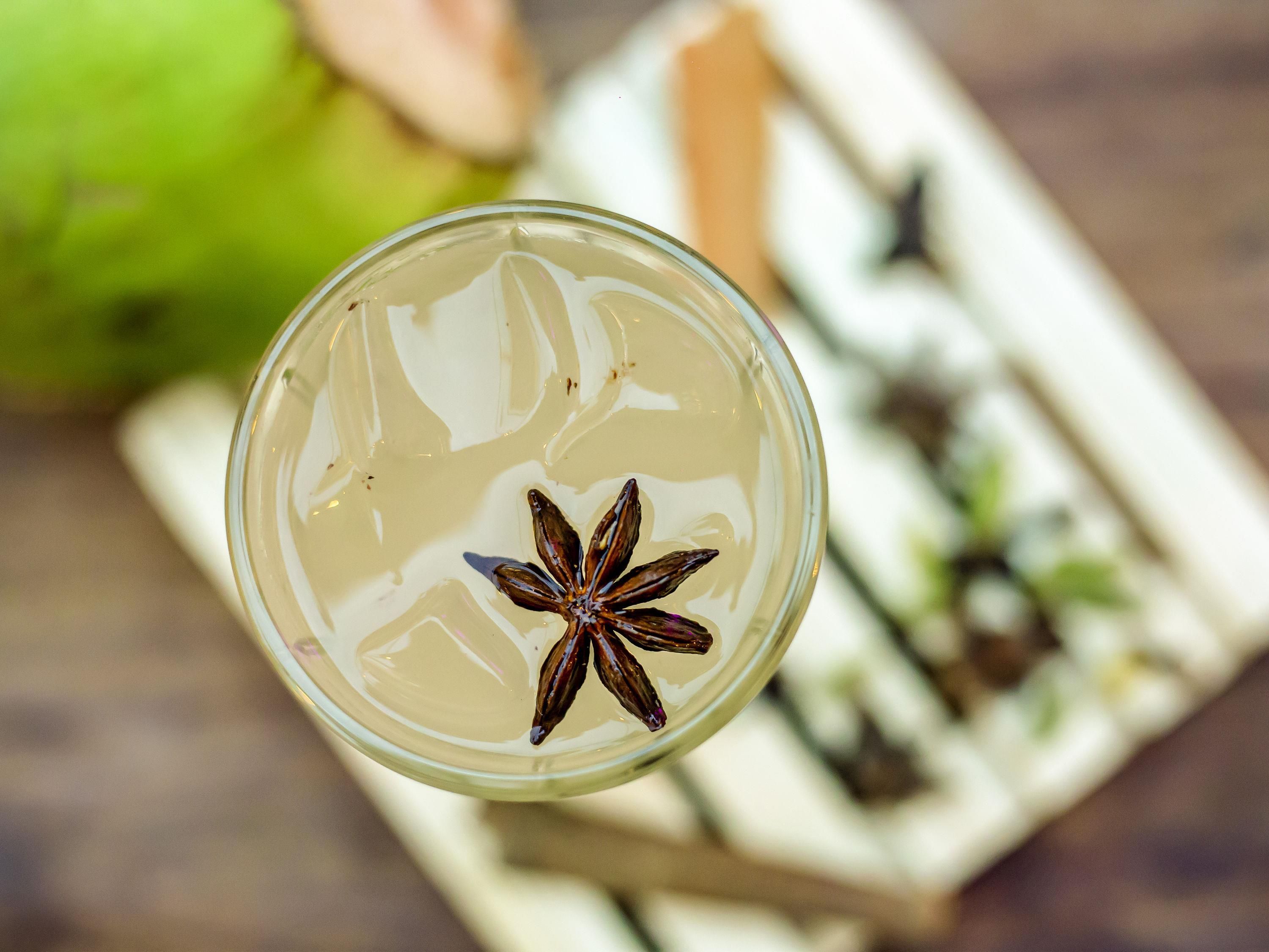 Un vaso con una estrella de anís flotando en un líquido transparente, sobre un fondo desenfocado que incluye un objeto verde y un objeto rectangular blanco con motas negras.
