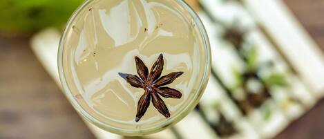Un vaso con una estrella de anís flotando en un líquido transparente, sobre un fondo desenfocado que incluye un objeto verde y un objeto rectangular blanco con motas negras.