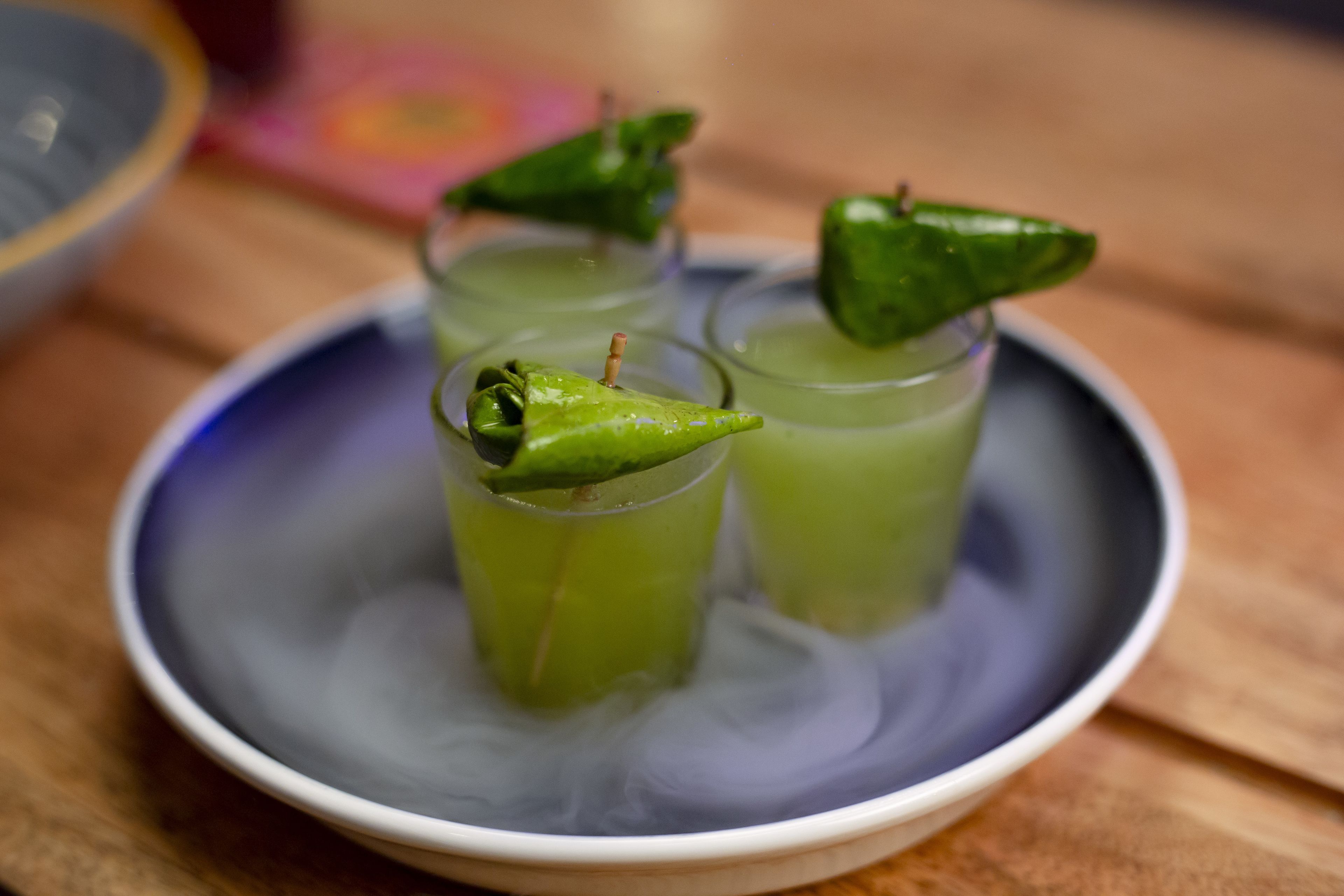 A bowl with three glasses of green liquid, each garnished with a green chili.