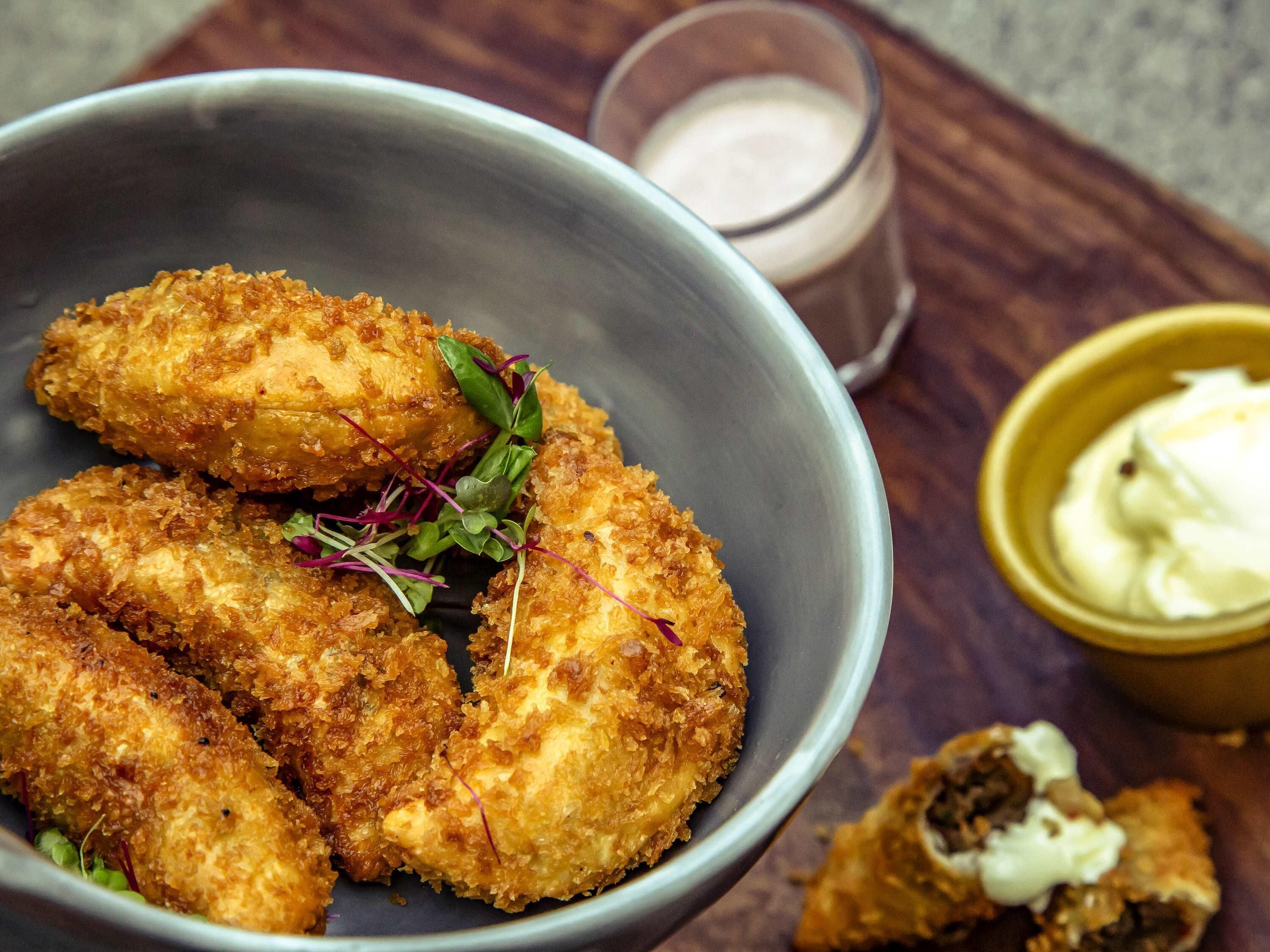 Un tazón de comida rebozada y frita, decorada con vegetales, acompañado de un vaso de una bebida cremosa y un pequeño tazón de salsa.