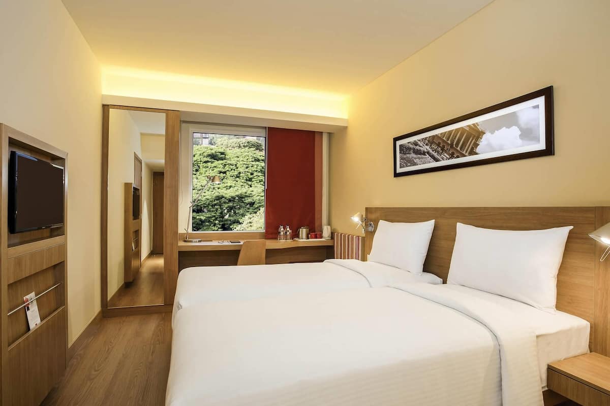 A hotel room with a large bed, a TV, a desk with a lamp, and a window with a view of greenery.