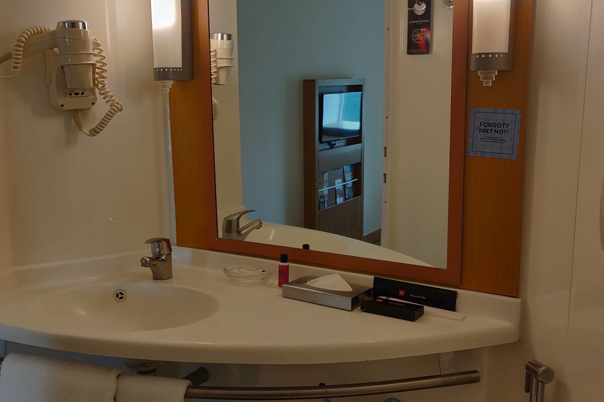 Un baño de hotel con un lavabo blanco, un espejo, un secador de pelo y un televisor al fondo.