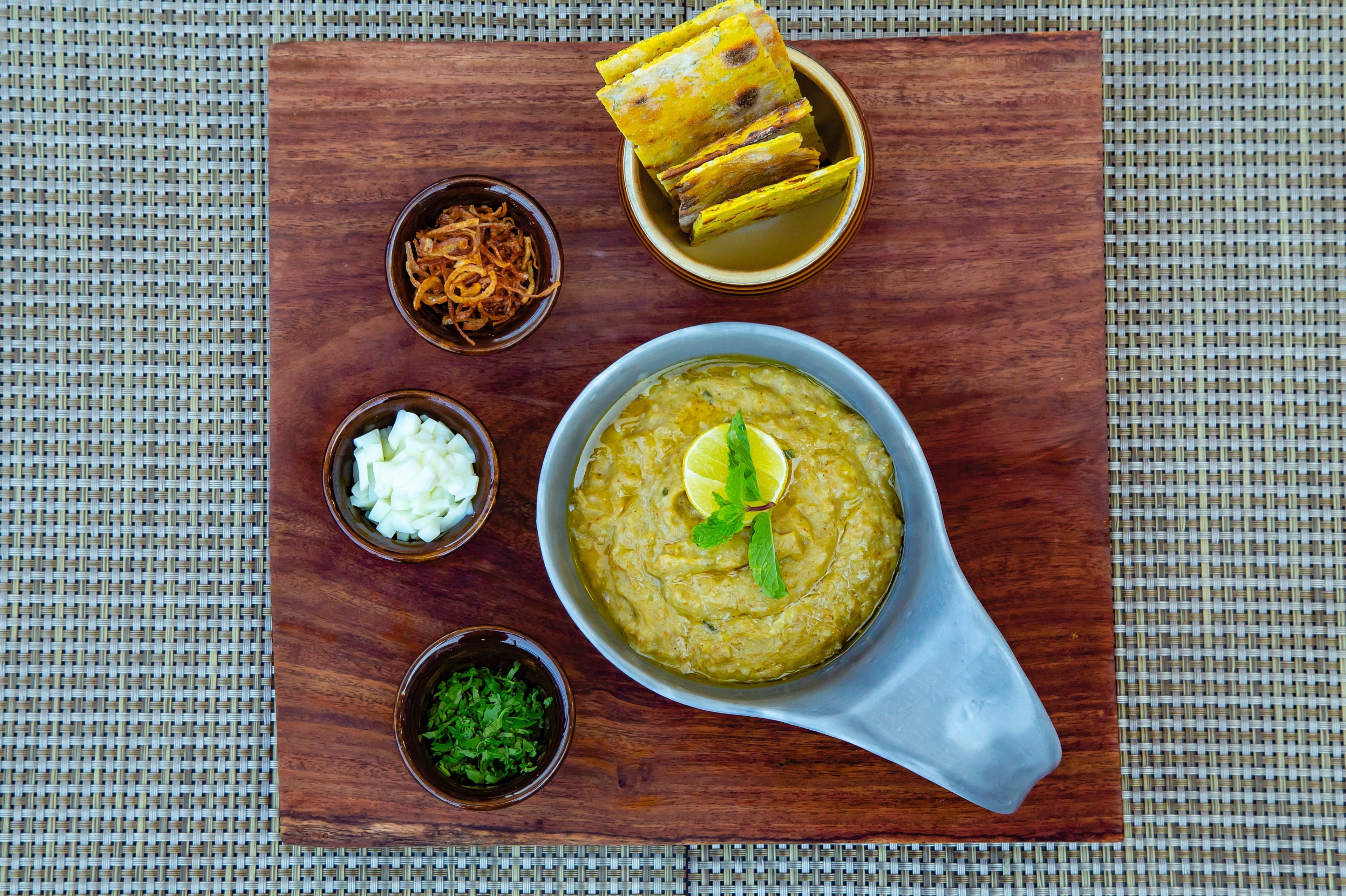A meal with a bowl of soup, a bowl of onions, a bowl of white sauce, a bowl of green garnish, and a stack of flatbreads on a wooden board.
