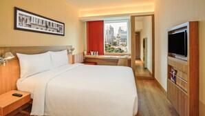 Habitación de hotel con una cama grande, televisión, escritorio con silla, ventana con vista a la ciudad y un cuadro en la pared.